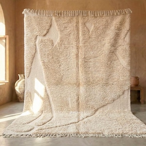Puede incluir: Una alfombra grande de color crema con un diseño abstracto texturizado. La alfombra presenta un pelo suave y afelpado y un borde con flecos. La alfombra se exhibe en una habitación con luz natural y una paleta de colores neutros.