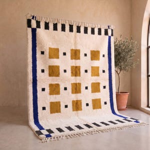 Puede incluir: Una alfombra rectangular con base color blanco roto que presenta una cuadrícula de cuadrados color amarillo mostaza y pequeños cuadrados negros. La alfombra está enmarcada por gruesas franjas azules verticales y bordes a cuadros negros y blancos con flecos.