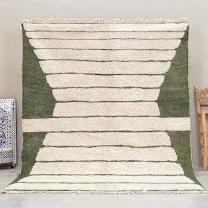 May include: A large, rectangular rug with a cream-colored background and horizontal stripes. The stripes are a muted green color. The rug has a fringe along the bottom edge. A decorative mosaic tile box and a wooden chair are in the background.