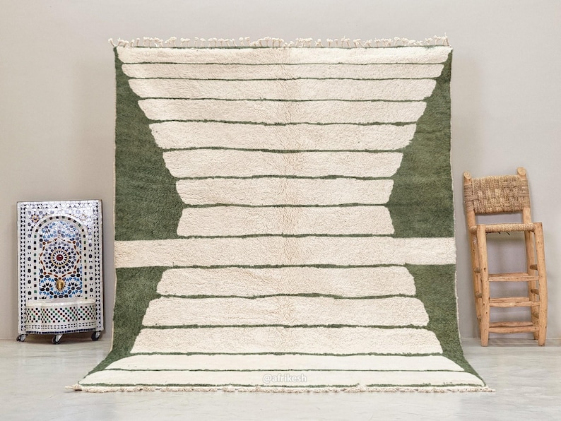 May include: A large, rectangular rug with a cream-colored background and horizontal stripes. The stripes are a muted green color. The rug has a fringe along the bottom edge. A decorative mosaic tile box and a wooden chair are in the background.