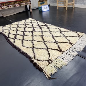 May include: A white and brown Moroccan rug with a diamond pattern and fringe. The rug is laid out on a dark floor.