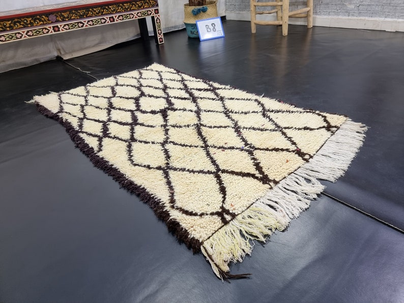 May include: A white and brown Moroccan rug with a diamond pattern and fringe. The rug is laid out on a dark floor.