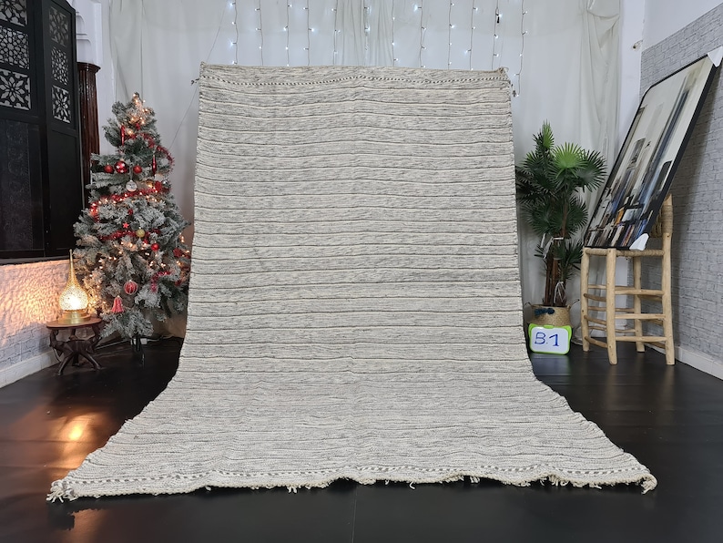 Puede incluir: Una alfombra grande, rectangular, de rayas blancas y grises con un borde de flecos. La alfombra est&aacute; colocada sobre un suelo oscuro en una habitaci&oacute;n con un &aacute;rbol de Navidad y otras decoraciones.