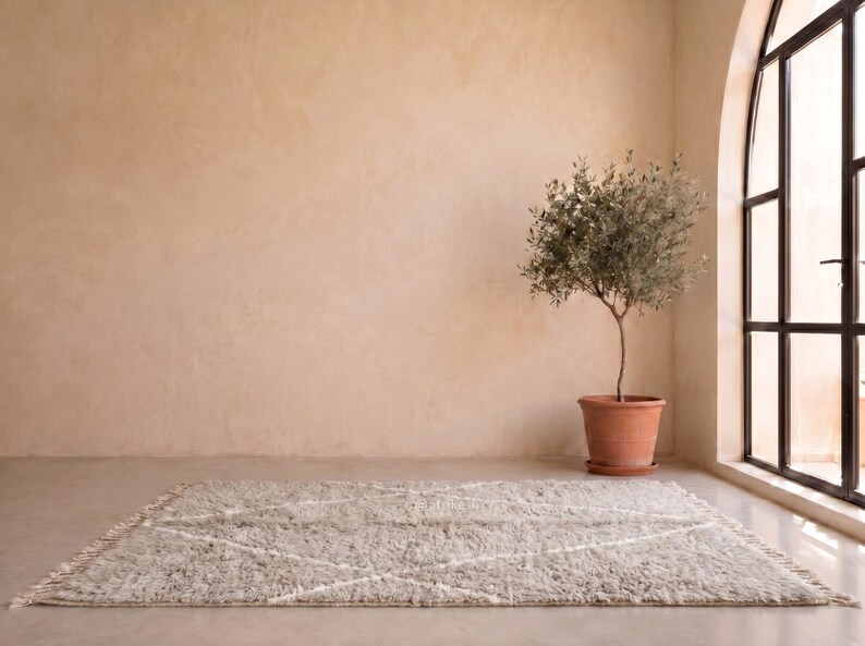 May include: A rectangular area rug featuring a shaggy, textured pile in a light beige tone with a subtle white diamond pattern and fringed edges on the short sides, positioned flat on a smooth floor surface in a room with a potted olive tree.