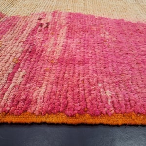 May include: Close-up of a pink and orange woven rug with a textured surface. The rug has a fringe border.