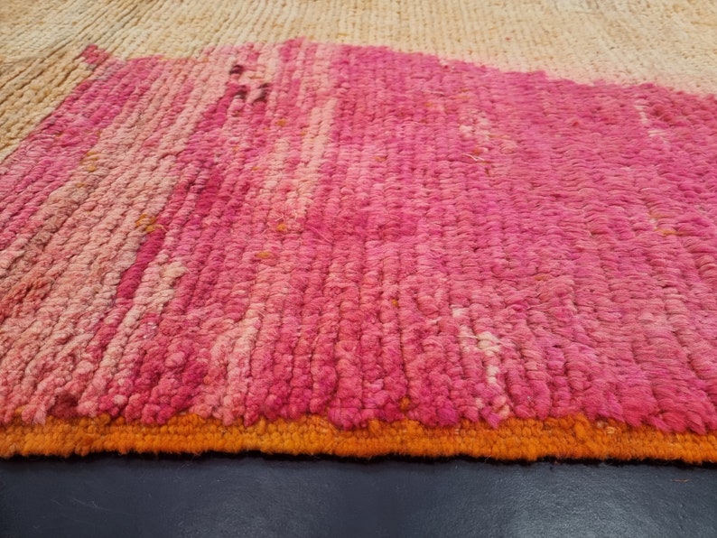 May include: Close-up of a pink and orange woven rug with a textured surface. The rug has a fringe border.