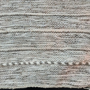 Puede incluir: Primer plano de una alfombra tejida con un patr&oacute;n de rayas blancas y grises. La alfombra tiene una superficie texturizada y un brillo sutil.
