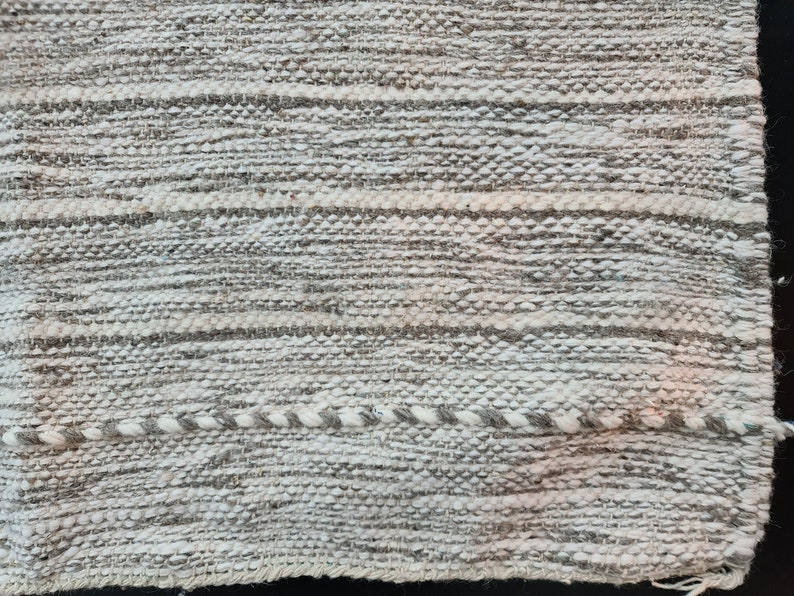 Puede incluir: Primer plano de una alfombra tejida con un patr&oacute;n de rayas blancas y grises. La alfombra tiene una superficie texturizada y un brillo sutil.
