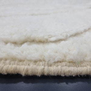 May include: Close-up of the edge of a white and beige shag rug. The rug has a thick, fluffy texture and a woven edge.