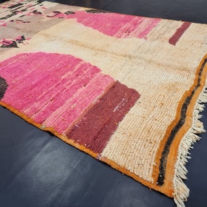 May include: A hand-woven rug with a colorful geometric pattern. The rug is made of wool and has a fringe border. The colors in the rug are pink, orange, brown, and black.