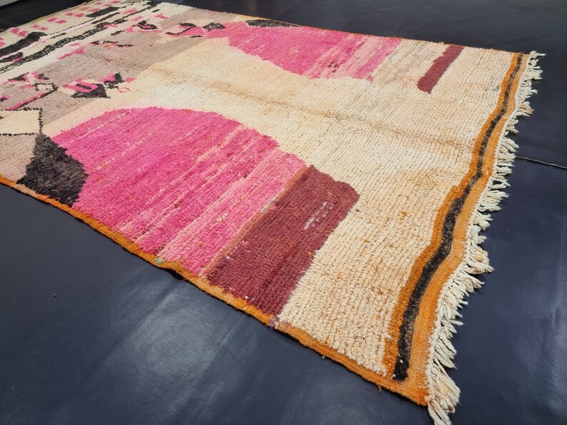 May include: A hand-woven rug with a colorful geometric pattern. The rug is made of wool and has a fringe border. The colors in the rug are pink, orange, brown, and black.