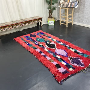 May include: A red, blue, green, and purple Moroccan rug with a geometric pattern. The rug is on a black floor in front of a wooden bench with a black and gold painted design.