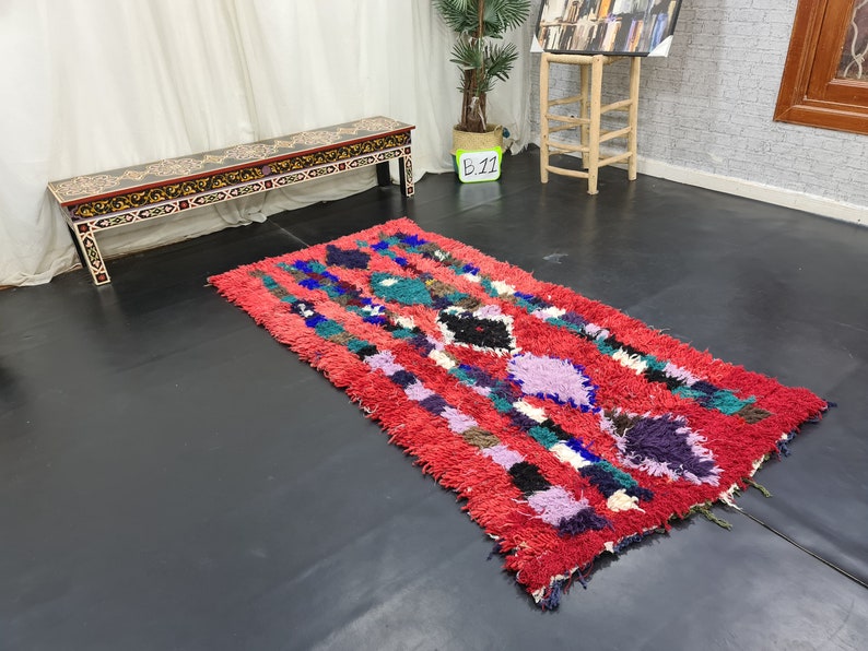 May include: A red, blue, green, and purple Moroccan rug with a geometric pattern. The rug is on a black floor in front of a wooden bench with a black and gold painted design.