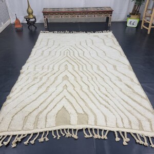 May include: A white and cream patterned rug with a geometric design. The rug has a fringe border and is made of a soft, fluffy material.