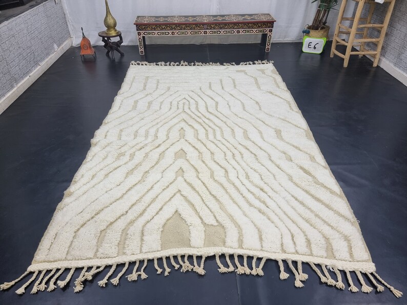 May include: A white and cream patterned rug with a geometric design. The rug has a fringe border and is made of a soft, fluffy material.