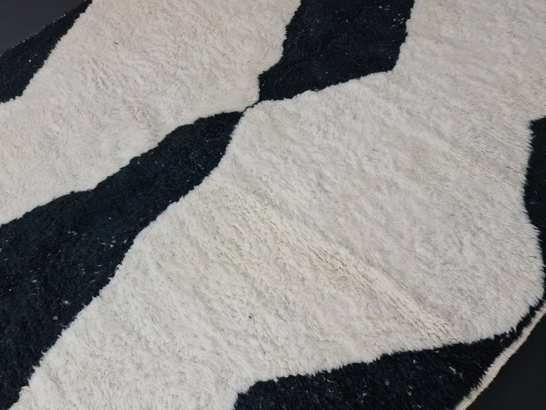 May include: Black and white geometric patterned rug with a fluffy texture. The rug has a wavy design with black and white stripes.