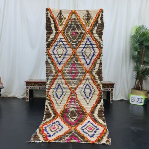 May include: A long, rectangular, white, brown, blue, pink, and orange Moroccan rug with a diamond pattern. The rug is hanging on a white wall.