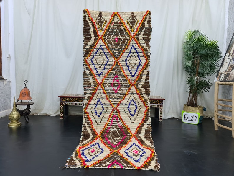 May include: A long, rectangular, white, brown, blue, pink, and orange Moroccan rug with a diamond pattern. The rug is hanging on a white wall.