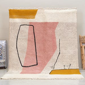 Puede incluir: Alfombra geométrica abstracta con fondo crema, con una gran forma rosa, líneas negras y acentos amarillo mostaza. La alfombra tiene una superficie texturizada y un borde con flecos. Una lámpara decorativa y una silla de madera están en el fondo.
