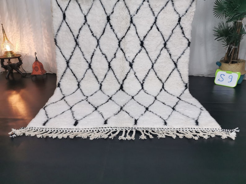 May include: White and black Moroccan rug with a diamond pattern and fringe. The rug is made of wool and is approximately 5 feet long.