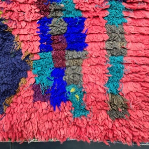 May include: A close-up of a colorful, hand-woven rug with a textured, fuzzy surface. The rug is made of different colored yarns, including red, blue, green, and brown. The rug has a geometric pattern.