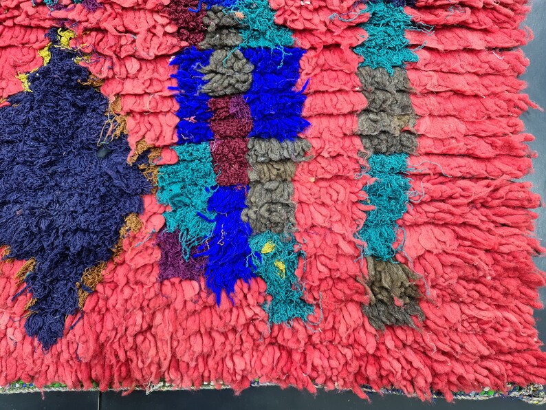 May include: A close-up of a colorful, hand-woven rug with a textured, fuzzy surface. The rug is made of different colored yarns, including red, blue, green, and brown. The rug has a geometric pattern.