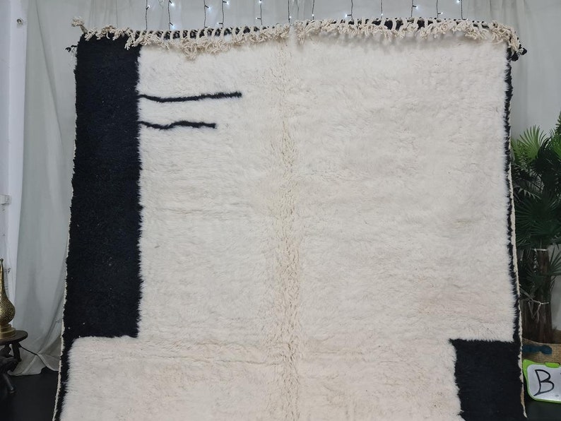 May include: A white and black Moroccan rug with a geometric design. The rug has a fringe border and is made of wool.