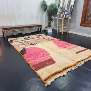 May include: A large, hand-woven rug with a cream background and abstract pink, brown, and black designs. The rug has a fringe border.