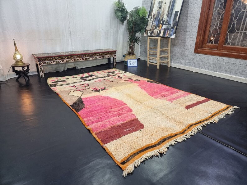 May include: A large, hand-woven rug with a cream background and abstract pink, brown, and black designs. The rug has a fringe border.