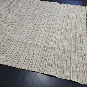 Puede incluir: Una alfombra tejida a rayas blancas y grises con un borde de flecos. La alfombra tiene una superficie texturizada y un brillo sutil.