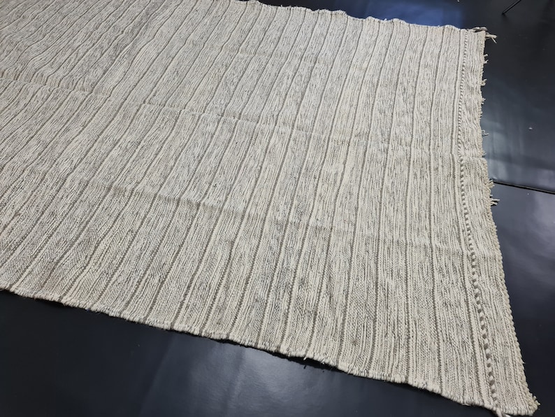 Puede incluir: Una alfombra tejida a rayas blancas y grises con un borde de flecos. La alfombra tiene una superficie texturizada y un brillo sutil.