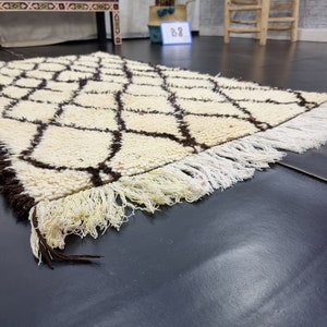 May include: A white and brown Moroccan rug with a geometric pattern and fringe. The rug is lying on a dark gray surface.