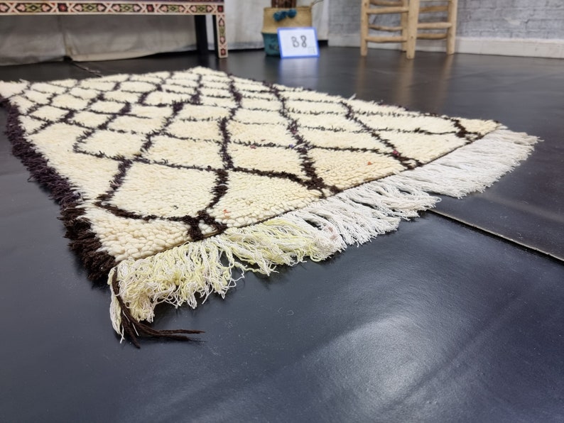 May include: A white and brown Moroccan rug with a geometric pattern and fringe. The rug is lying on a dark gray surface.