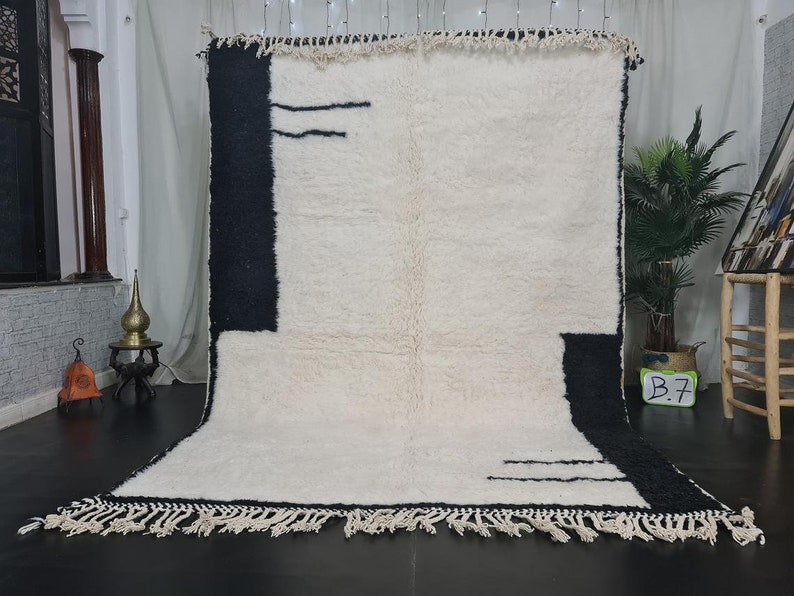 May include: A white and black Moroccan rug with a geometric design. The rug has a fringe border and is made of wool.