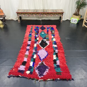 May include: A red, hand-woven rug with a colorful geometric pattern. The rug has a fringe border and is made of wool. The rug is approximately 8 feet long and 3 feet wide.