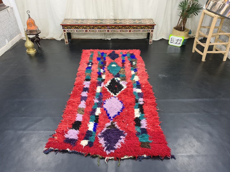 May include: A red, hand-woven rug with a colorful geometric pattern. The rug has a fringe border and is made of wool. The rug is approximately 8 feet long and 3 feet wide.