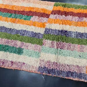 May include: A colorful striped rug with a variety of colors including orange, green, purple, and white. The rug is made of a soft, fluffy material.