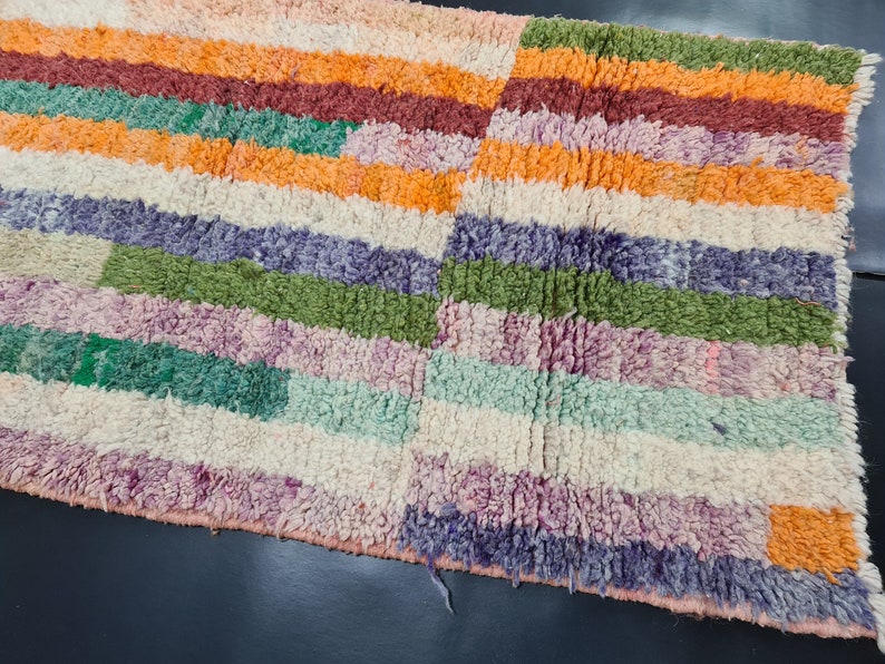 May include: A colorful striped rug with a variety of colors including orange, green, purple, and white. The rug is made of a soft, fluffy material.