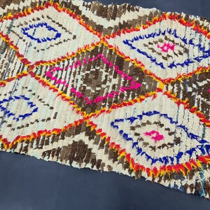 May include: A white, brown, blue, yellow, and pink Moroccan rug with a diamond pattern. The rug is handwoven and has a fringed edge.