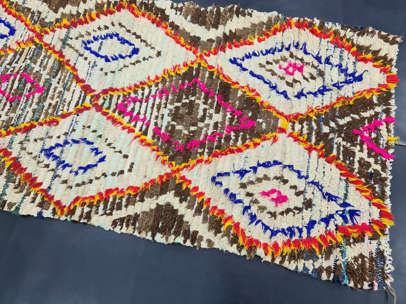 May include: A white, brown, blue, yellow, and pink Moroccan rug with a diamond pattern. The rug is handwoven and has a fringed edge.