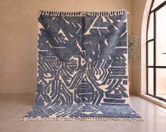 May include: A rectangular area rug featuring a deep blue abstract geometric pattern on a cream-colored background. The rug has a thick, textured pile and is finished with short, cream-colored fringe along the top and bottom edges.