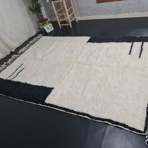 May include: A white and black geometric patterned rug with fringe on all sides. The rug has a modern design with a black border and a white centre.