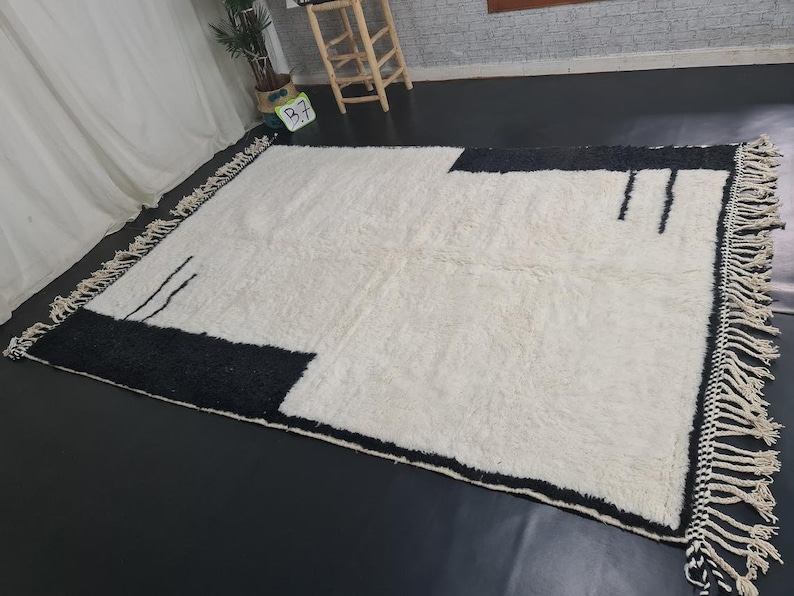 May include: A white and black geometric patterned rug with fringe on all sides. The rug has a modern design with a black border and a white centre.
