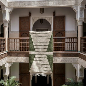 May include: A rectangular, handwoven rug with a geometric design, featuring alternating stripes of off-white and olive green. The rug is displayed in a courtyard with wooden balconies and carved architectural details, creating a warm, inviting atmosphere.