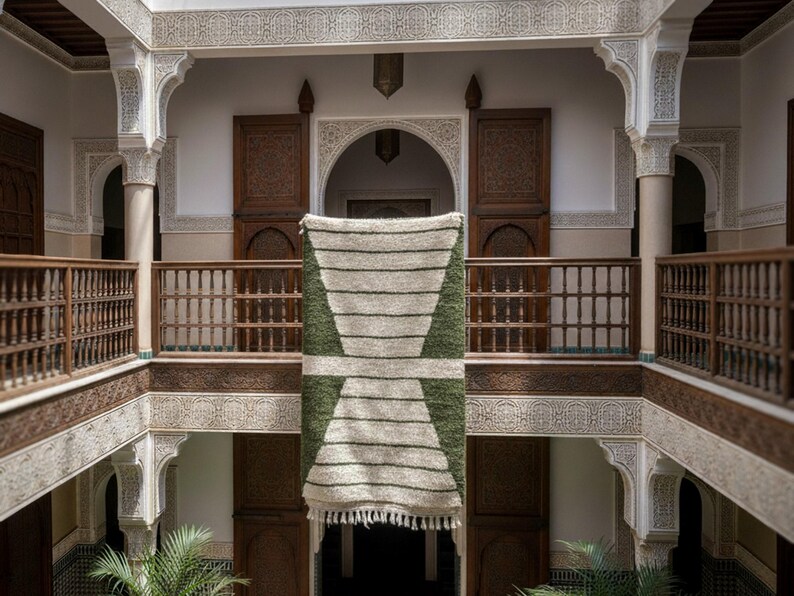 May include: A rectangular, handwoven rug with a geometric design, featuring alternating stripes of off-white and olive green. The rug is displayed in a courtyard with wooden balconies and carved architectural details, creating a warm, inviting atmosphere.