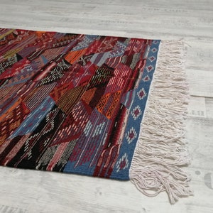 Pu&ograve; includere: Un tappeto colorato con motivi geometrici e un design patchwork. Il tappeto presenta una variet&agrave; di colori tra cui rosso, blu, arancione e nero. I bordi sono rifiniti con un bordo blu e frange color crema.