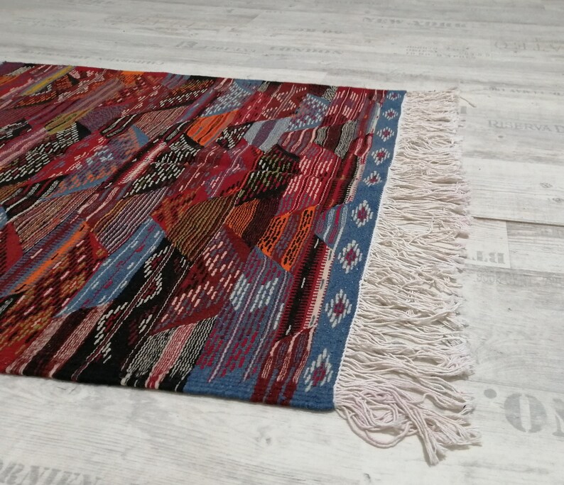Pu&ograve; includere: Un tappeto colorato con motivi geometrici e un design patchwork. Il tappeto presenta una variet&agrave; di colori tra cui rosso, blu, arancione e nero. I bordi sono rifiniti con un bordo blu e frange color crema.