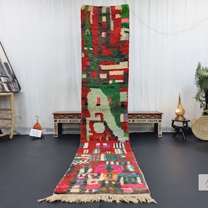 Pode incluir: Um tapete marroquino longo e colorido com um padrão geométrico em tons de vermelho, verde, preto e branco. O tapete tem franjas em ambas as extremidades e está colocado sobre um piso preto.