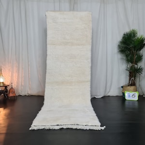 May include: A white shag rug with a fringe border. The rug is hanging on a wall in a room with white curtains.