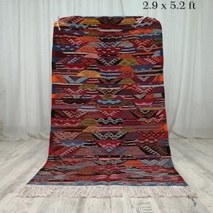 Puede incluir: Una alfombra rectangular tejida a mano con un patrón geométrico. La alfombra mide aproximadamente 88 x 158 cm y presenta una vibrante mezcla de colores, incluyendo rojo, naranja, azul y marrón. Tiene un borde con flecos y una superficie texturizada.